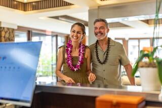 A smiling couple stands together at a reception desk, wearing floral leis and casual attire, showcasing a relaxed, tropical vibe.