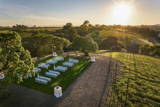 A picturesque outdoor venue features neatly arranged chairs on green grass, surrounded by vineyards under a warm, glowing sunset.