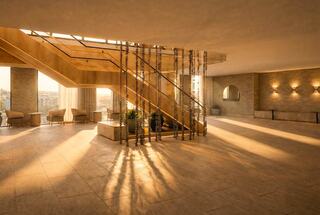 A spacious lobby features wooden stairs, soft lighting, and elegant decor. Sunlight casts long shadows, enhancing the serene atmosphere.