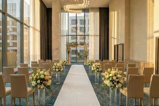 A beautifully decorated wedding venue features a floral arch, elegant chairs, and a white aisle, illuminated by natural light.