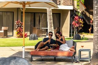 A couple enjoys a relaxed outdoor meal, surrounded by tropical scenery, sipping drinks and soaking up the sun under a beach umbrella.