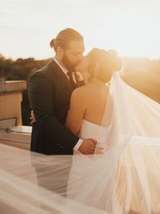 A couple shares a romantic kiss at sunset, surrounded by flowing wedding veil, embodying love and warmth in a beautiful moment.