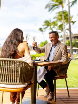 A couple enjoys a celebratory toast outdoors, surrounded by palm trees, seated at a stylish table, exuding a joyful, relaxed atmosphere.