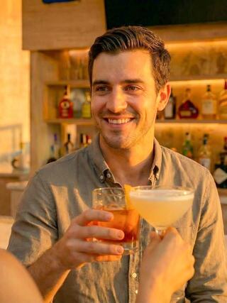 A smiling man holds two cocktails, enjoying a lively moment at a bar with a well-stocked shelf of drinks in the background.