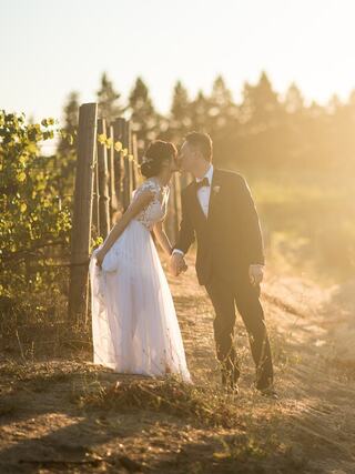 A couple in formal attire shares a romantic kiss in a sunlit vineyard, surrounded by nature's beauty and soft golden light.
