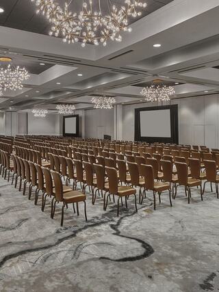 A spacious conference room filled with rows of brown chairs, elegant lighting fixtures, and two large screens at the front.