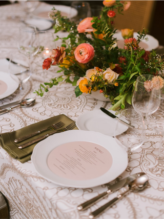 A beautifully set table features elegant plates, cutlery, and a vibrant floral centerpiece, all atop an intricately patterned cloth.