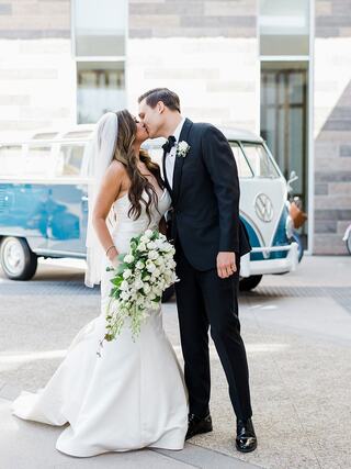 A couple shares a romantic kiss in wedding attire, surrounded by modern architecture and a vintage blue VW bus in the background.