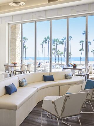 A bright, modern lounge features curved seating with blue pillows and large windows showcasing palm trees and ocean views.
