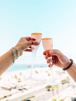 Two hands raise elegant glasses filled with pink bubbly, celebrating a sunny day by the beach, with a clear sky and distant ocean view.