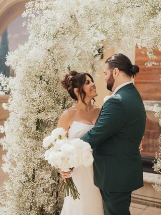 A couple shares a tender moment under a floral arch, surrounded by delicate white blooms, radiating love and joy on their special day.