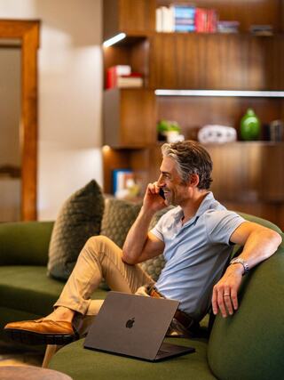 A man in casual attire sits on a green sofa, talking on the phone with a laptop in front of him, surrounded by stylish decor.