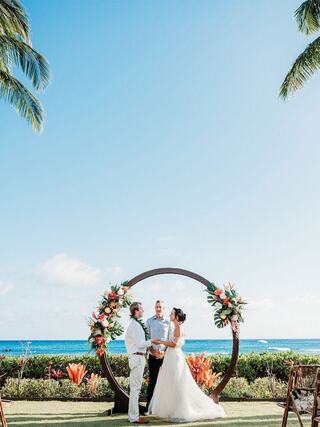 A couple exchanges vows under a floral arch with the ocean backdrop, surrounded by lush greenery and blue skies, creating a romantic atmosphere.