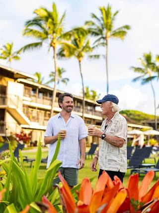 Two men enjoying a sunny day in a tropical setting, laughing and holding drinks, surrounded by vibrant flowers and palm trees.