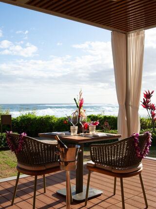 A cozy dining setup overlooks a scenic ocean view, accented by vibrant tropical flowers and soft, flowing curtains.