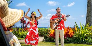 A man plays guitar while a woman dances joyfully in vibrant floral outfits, against a tropical backdrop of lush greenery and ocean.