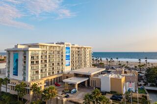 A modern hotel overlooks a sandy beach, surrounded by palm trees, with a clear sky and ocean in the background, embodying a tropical escape.