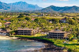 Lush coastal landscape featuring low-rise buildings surrounded by palm trees, with mountains in the background and gentle waves lapping the shore.
