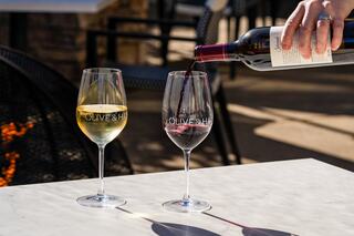 A hand pours red wine into a glass beside a white wine glass, both featuring "OLIVE & H" etched on them, set on a marble table.