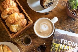 A cozy scene featuring croissants, a cappuccino with latte art, and a book on Napa, alongside a small dessert and succulent plant.