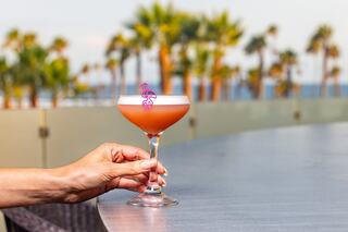 A hand holds a vibrant cocktail with a pink flamingo stirrer, set against a backdrop of palm trees and a sunny sky.
