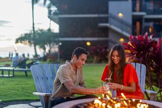 A couple enjoys drinks by a fire pit, laughing and sharing a moment in a vibrant outdoor setting with lush greenery at sunset.