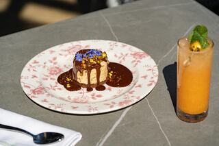 A beautifully plated dessert topped with flowers and nuts sits in chocolate sauce, accompanied by a vibrant orange drink and a mint garnish.