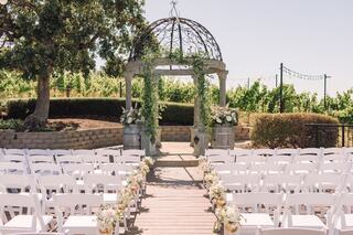 A charming outdoor wedding venue features a floral-adorned gazebo surrounded by rows of white chairs, set against a vineyard backdrop.