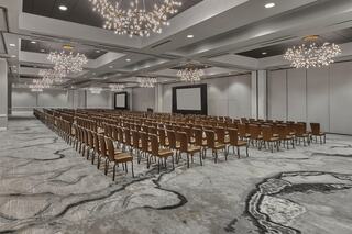 A spacious, modern conference room featuring rows of brown chairs, elegant chandeliers, and a large screen at the front.