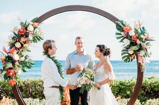 A couple stands before a celebrant under a floral arch, exchanging vows against a backdrop of ocean waves and a clear blue sky.