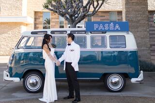 A couple stands hand in hand by a vintage blue van, dressed elegantly for a special occasion, radiating romance and joy.