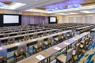 A spacious conference room with rows of tables and chairs, equipped for presentations, featuring screens and a modern design.