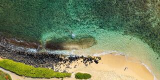 Crystal-clear turquoise waters meet a sandy beach, with rocky patches and lush green vegetation lining the shore, creating a serene tropical scene.