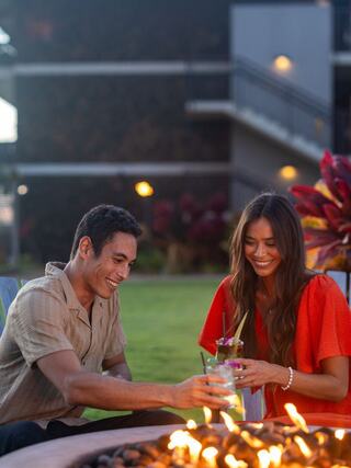 Two smiling individuals enjoy drinks while seated by a fire pit in a cozy outdoor setting, surrounded by vibrant plants and evening lights.