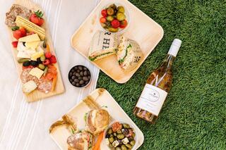 A picnic spread features cheese, fruits, sandwiches, olives, and wine, set on green grass, evoking a relaxing outdoor dining vibe.